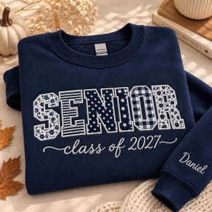 May include: A navy blue crewneck sweatshirt featuring the word SENIOR in large block letters with varied patterns including floral, stripes, polka dots, stars, and gingham. Below is the text class of 2027, and the name Daniel is on the sleeve.