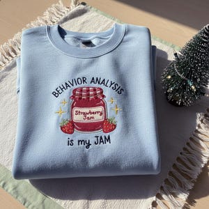 以下が含まれることがあります： 水色のスウェットシャツ。刺繍のデザインには、イチゴジャムの瓶、「BEHAVIOR ANALYSIS」と「is my JAM」の文字、2つのイチゴ、星のアクセントが含まれています。
