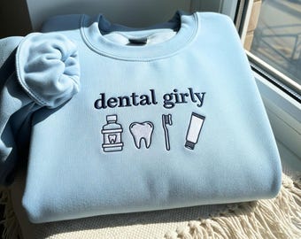 May include: Light blue sweatshirt with the embroidered text "dental girly" and dental-themed icons. The design includes a mouthwash bottle, a tooth, a toothbrush, and a tube of toothpaste. The sweatshirt is folded on a cream-colored surface.