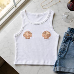 May include: White ribbed vest top featuring two embroidered seashell designs. The top has a scoop neck and is sleeveless. A pair of denim shorts and a glass of red wine are also in the image. A gold necklace is also visible.