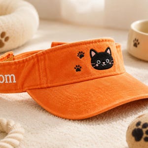 May include: Bright orange visor featuring a black cat face and paw prints. The word "tom" is embroidered on the band. A pet-themed accessory.