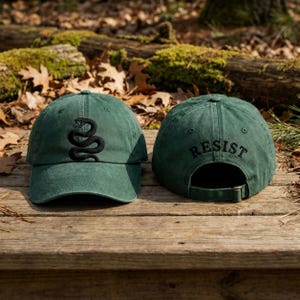 May include: Two forest green baseball caps made of a washed cotton fabric. The front cap features an embroidered black snake graphic. The back cap shows the word RESIST embroidered in black above the adjustable strap with a metal buckle closure.