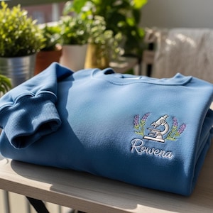 Puede incluir: Sudadera azul claro con un diseño bordado detallado. El diseño presenta un microscopio, ramitas de lavanda y el nombre "Rowena" en escritura blanca. La sudadera está doblada sobre una superficie de madera.