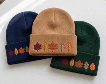 May include: Three knit beanies in navy blue, tan, and dark green. Each hat features a folded cuff embroidered with a row of five colorful autumn leaves in shades of burgundy, yellow, orange, and light brown, arranged in a repeating pattern.