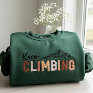 May include: A dark green sweatshirt with the embroidered phrase "Keep CLIMBING" in black, peach, and white. The design includes a mountain range graphic. The sweatshirt is folded and displayed near a window with a vase of white flowers.