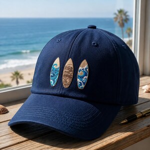 May include: A navy blue baseball cap featuring three embroidered surfboard-shaped patches on the front. The outer two patches display blue and white wave patterns, while the centre patch features a detailed brown and tan line art wave design.