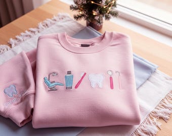 May include: A light pink sweatshirt and sleeve with embroidered dental-themed designs. The sweatshirt features a dental chair, toothpaste, toothbrush, tooth, and dental tools. The sleeve has a tooth and the name "Sarah, RDH".
