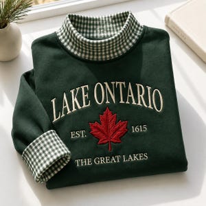 Può includere: Una felpa a maniche lunghe verde scuro con collo alto a motivo vichy verde e bianco. La parte anteriore presenta il testo ricamato LAKE ONTARIO, EST. 1615 e THE GREAT LAKES, centrato con una grafica di una foglia d'acero rossa.