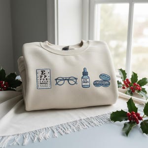 May include: A cream-colored sweatshirt embroidered with eye care symbols. The design includes a vision chart, eyeglasses, a bottle of eye drops, and contact lenses, all in blue thread. The sweatshirt is folded on a white surface.