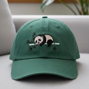 Può includere: Cappellino da baseball verde con ricamo di un panda che dorme su un ramo di bambù. Il panda è nero e bianco, con il bambù e le foglie nere. Il cappellino ha una visiera curva e una chiusura in tessuto.
