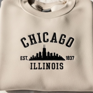 Op de afbeelding: Een beige sweatshirt met het woord "CHICAGO" in zwart geborduurd, boven een zwart silhouet van de skyline. Daaronder de woorden "EST. 1837" en "ILLINOIS". De sweatshirt is gemaakt van zacht materiaal.