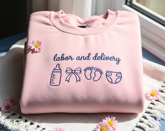 Op de afbeelding: Een lichtroze sweatshirt met de woorden "labor and delivery" in marineblauw geborduurd. Onder de tekst staan marineblauwe borduursels van een babyfles, een strik, voetafdrukken en een luier. Het sweatshirt ligt op een witte doek met franjes.