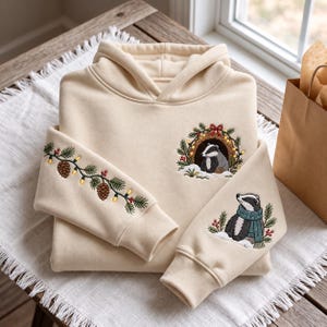 May include: A cream-colored hooded sweatshirt featuring detailed embroidery. The chest displays a badger inside a festive wreath. One sleeve shows a pine branch with pinecones and string lights, while the other features a badger wearing a teal scarf.