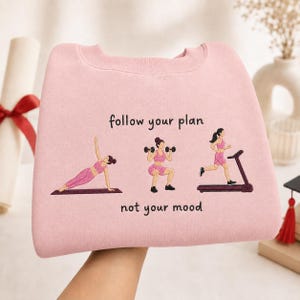 May include: A light pink sweatshirt featuring embroidered illustrations of a person exercising in three different ways: doing a side plank, lifting dumbbells, and running on a treadmill. The text "follow your plan" and "not your mood" is embroidered.