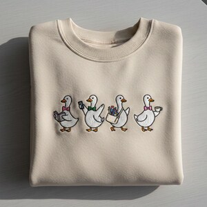 Può includere: Felpa girocollo beige con un design ricamato di quattro oche dei cartoni animati. Ogni oca è raffigurata con un accessorio diverso: un libro, un telefono, una borsa di provviste e una tazza di caffè. Le oche indossano papillon di vari colori.