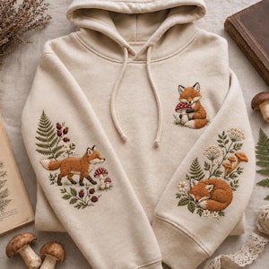 May include: A beige hooded sweatshirt featuring detailed embroidery of foxes, mushrooms, and botanical elements. The chest displays a small fox with a mushroom, while each sleeve showcases larger embroidered scenes of foxes in nature.