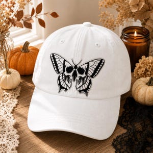 May include: A white baseball cap featuring a black and white embroidered graphic of a butterfly with a skull pattern on its wings. The cap has a structured crown, a curved brim, and a button on top, showcasing a detailed illustrative design.