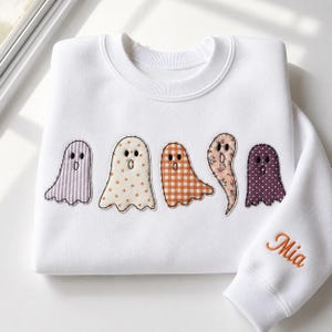 May include: A white crewneck sweatshirt featuring five ghost-shaped fabric appliqués across the chest, each with a unique pattern including stripes, stars, gingham, floral, and polka dots. The name Mia is embroidered in orange on the sleeve.