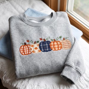 Puede incluir: Una sudadera gris jaspeado de cuello redondo con una fila bordada de cinco calabazas en el pecho. Las calabazas presentan varios patrones, incluyendo cuadros vichy naranjas, crema con pequeñas calabazas naranjas, cuadros azul marino, cuadros naranjas y rayas beige.