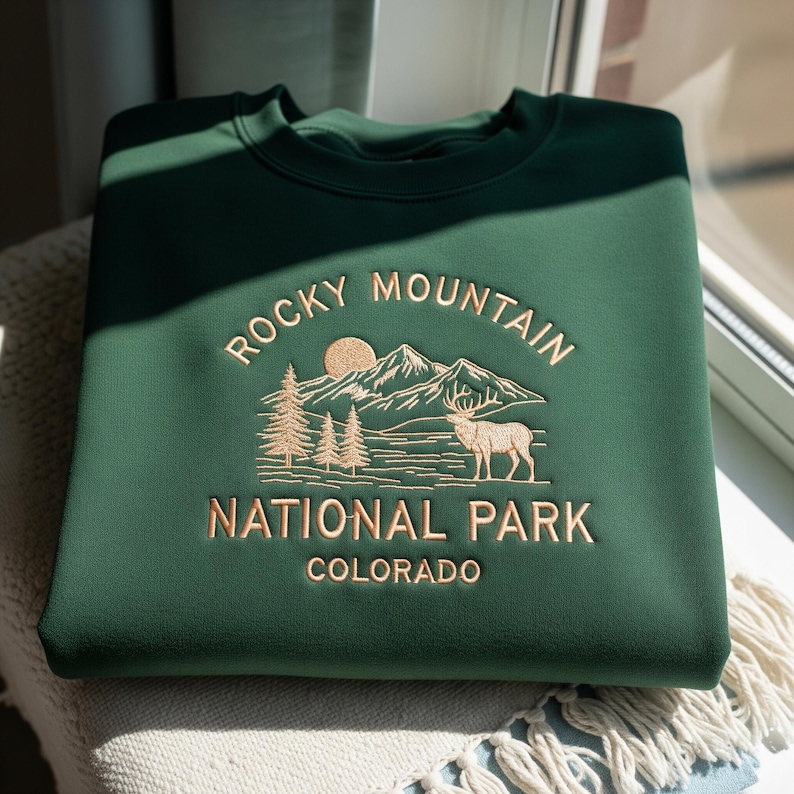 May include: A dark green sweatshirt with the embroidered text "Rocky Mountain National Park Colorado". The design features a mountain range, trees, a sun, and a deer in a tan color. The sweatshirt is folded on a light-colored surface.