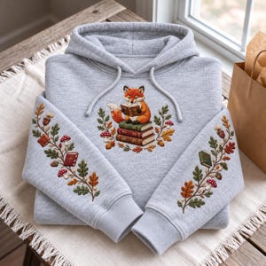 May include: A light gray hooded sweatshirt featuring an embroidered orange fox reading a book while sitting on a stack of three books. The sleeves and chest are decorated with embroidered oak leaves, acorns, and red-capped mushrooms.