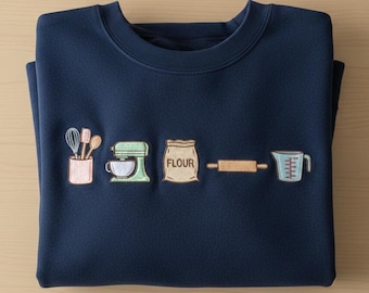 May include: Navy blue sweatshirt with embroidered baking-themed icons. The design includes a whisk, spatula, mixer, bag of flour, rolling pin, and measuring cup. The sweatshirt is folded neatly on a light wood surface.