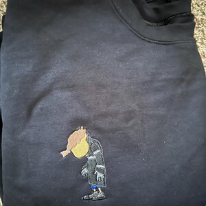 May include: Black sweatshirt with an embroidered cartoon character wearing a coat and hat. The character is yellow with a red head and is looking down.