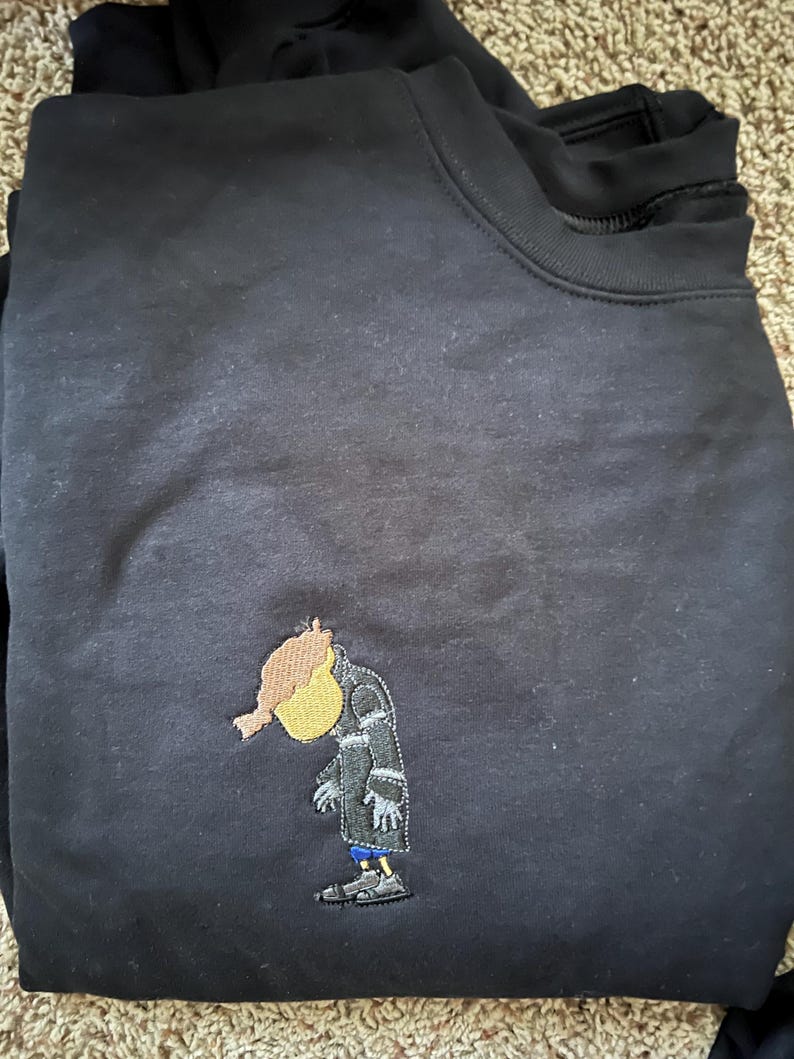 May include: Black sweatshirt with an embroidered cartoon character wearing a coat and hat. The character is yellow with a red head and is looking down.
