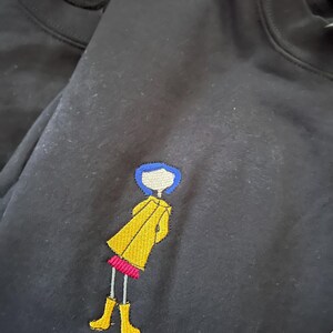May include: Black t-shirt with an embroidered image of a girl with blue hair wearing a yellow raincoat and red shoes.