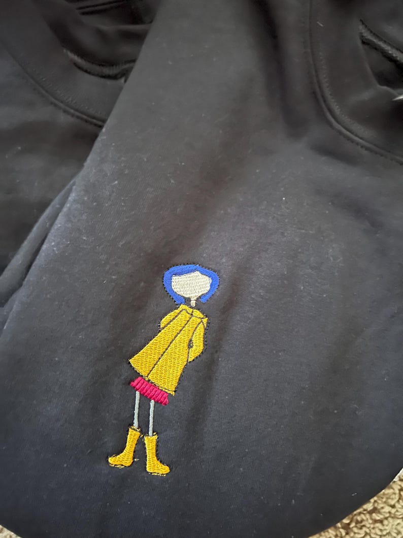 May include: Black t-shirt with an embroidered image of a girl with blue hair wearing a yellow raincoat and red shoes.