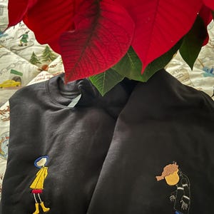 May include: Two black sweatshirts with embroidered images. One sweatshirt features a girl in a yellow raincoat and the other features a man in a black coat.