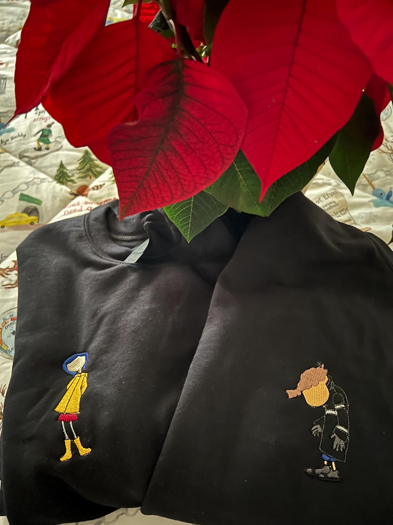 May include: Two black sweatshirts with embroidered images. One sweatshirt features a girl in a yellow raincoat and the other features a man in a black coat.