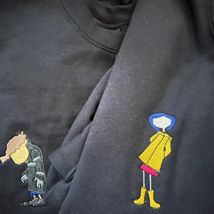 May include: Two black sweatshirts with embroidered images. One sweatshirt has a yellow raincoat-wearing girl with blue hair and the other has a brown-haired boy wearing a black coat.