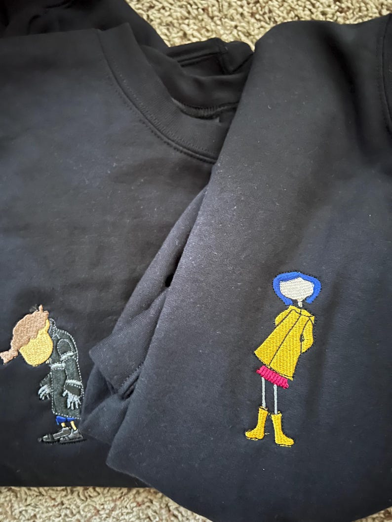 May include: Two black sweatshirts with embroidered images. One sweatshirt has a yellow raincoat-wearing girl with blue hair and the other has a brown-haired boy wearing a black coat.