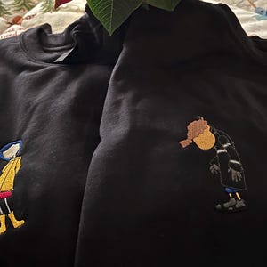 May include: Two black sweatshirts with embroidered images of cartoon characters. One sweatshirt features a girl in a yellow raincoat and red skirt, while the other sweatshirt features a person in a black coat and white scarf.