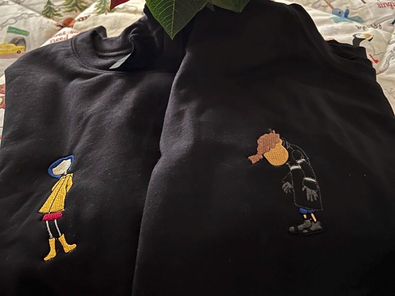 May include: Two black sweatshirts with embroidered images of cartoon characters. One sweatshirt features a girl in a yellow raincoat and red skirt, while the other sweatshirt features a person in a black coat and white scarf.