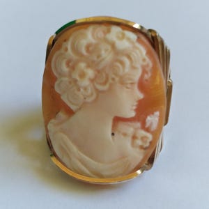 May include: Oval cameo ring featuring a carved profile of a woman with curly hair. The cameo is set in a gold-colored metal frame. The background of the cameo is orange, and the woman's profile is a creamy white color.