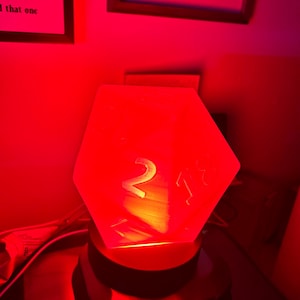 May include: A translucent red 20-sided die lamp resting on a circular black base. The die features large, embossed numbers including a prominent 2 and an 18, illuminated from within by a bright red light that casts a glow on the surrounding area.