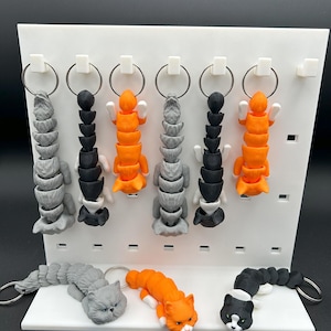 May include: A set of 3D printed cat keychains in various colors, including gray, black, and orange. The keychains are designed to look like cats with their tails curled up. The keychains are displayed on a white pegboard.