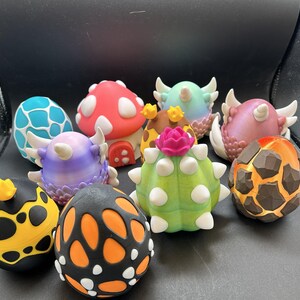 May include: A collection of ten small, egg-shaped figurines featuring various designs, including a red mushroom house, a green cactus with a pink flower, a blue and white cracked egg, a black and yellow spotted egg, and several dragon-like eggs.