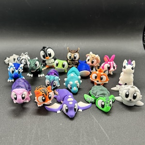 May include: A collection of small, multi-colored 3D printed animal figurines including a purple dragon, green turtle, blue triceratops, orange fox, white unicorn, black and white penguin, brown owl, and various other creatures with large eyes.