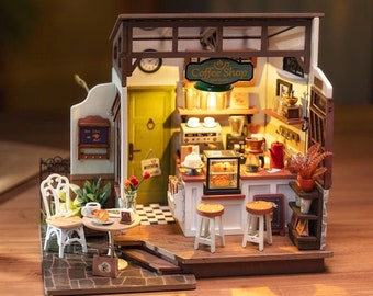 May include: A miniature dollhouse coffee shop with a green sign that reads "Coffee Shop" and a white counter with pastries and a coffee maker. There are two white chairs and a table with a pastry on it outside the shop.