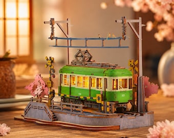May include: A wooden model of a green and cream-colored tram on tracks, featuring illuminated windows, overhead power lines with small bird figures, and decorative pink cherry blossom tree cutouts on a base with a railroad crossing sign.
