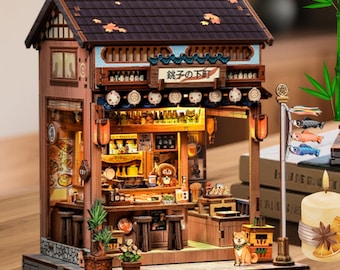 May include: A detailed wooden miniature of a Japanese shop with a tiled roof, lanterns, and the text "銚子の下町". The interior is lit, showcasing miniature bottles, tables, and a small dog figurine. A candle and bamboo stalks are in the foreground.