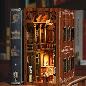 Puede incluir: Un rincón de libro de madera con un interior detallado que representa una escena de biblioteca. El exterior del rincón del libro es marrón con el texto "The Philosophical Library". Un libro azul está a la izquierda.