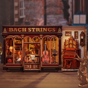 Può includere: Modello in legno dettagliato di un negozio di musica, con la scritta "BACH STRINGS" sopra l'ingresso. Il negozio espone vari strumenti a corda, tra cui violini e un violoncello. Una sezione separata mostra un organo e una statua.