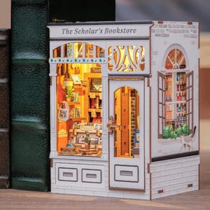 May include: A detailed miniature wooden model of a bookstore, labelled "The Scholar's Bookstore". The model has a white brick facade, a detailed interior with books and shelves, and a lit interior, creating a warm glow.