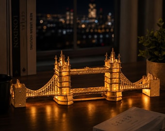 May include: A miniature wooden model of Tower Bridge featuring intricate laser-cut details and warm internal lighting that illuminates the structure. The model displays two main towers connected by a central walkway, resting on a flat surface.