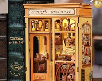 May include: A miniature wooden "Cooking Bookstore" model with detailed interior, lit windows, and a sign. Next to it, a green book with "Cooking Time" and a chef's hat and utensils. A golden Christmas ornament hangs in the background.