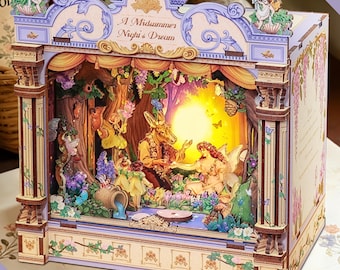 May include: A wooden diorama of a theater stage with the text "A Midsummer Night's Dream". The stage features a forest scene with fairies, a rabbit, and a glowing sun. The diorama is painted in pastel colors.